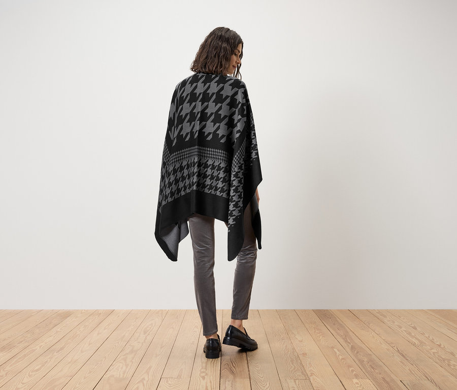 Nő visel fekete-szürke, kakasmintás poncsót, szürke nadrágot és fekete cipőt.
