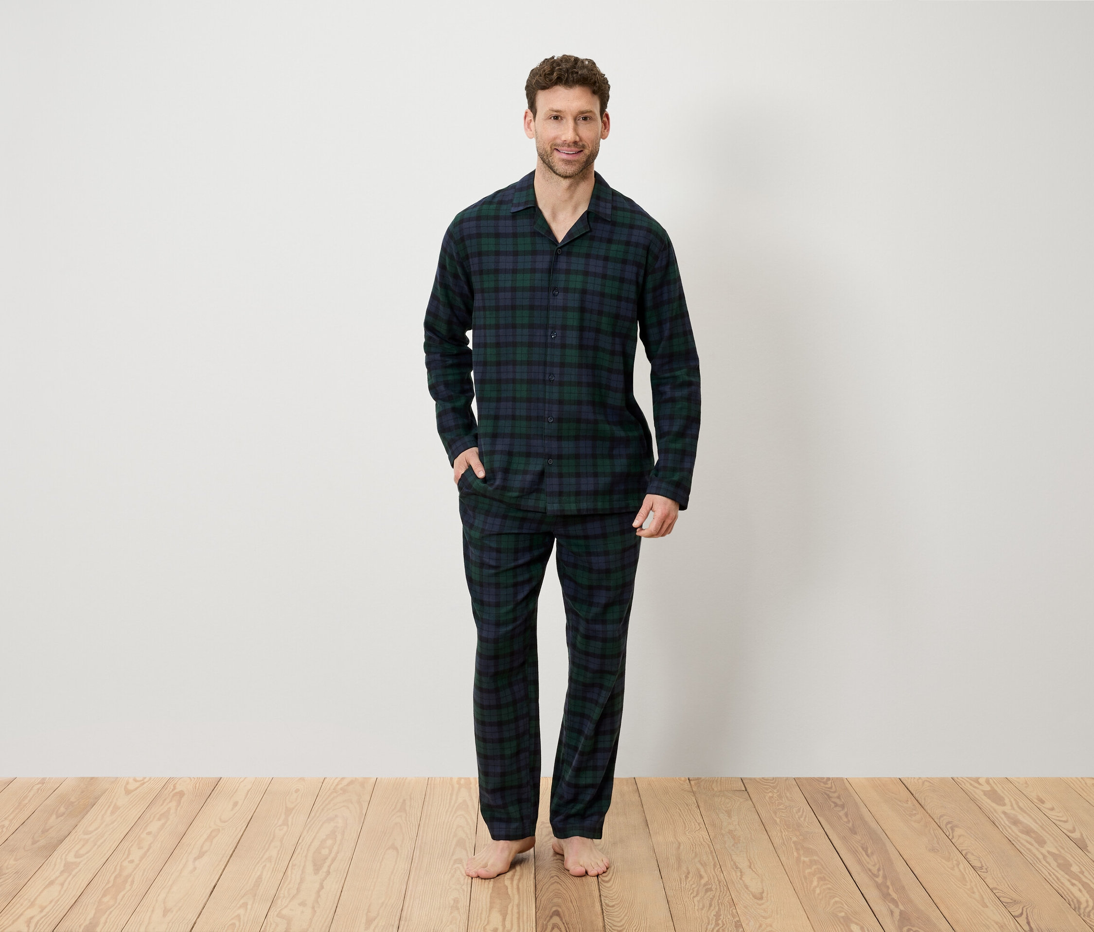 Egy kockás pizsamában lévő férfi áll egy fapadlón egy fehér fal előtt.