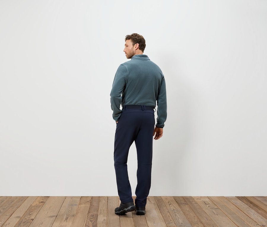 Férfi kék nadrágban és polár pulóverben áll háttal a kamerának egy fapadlón.