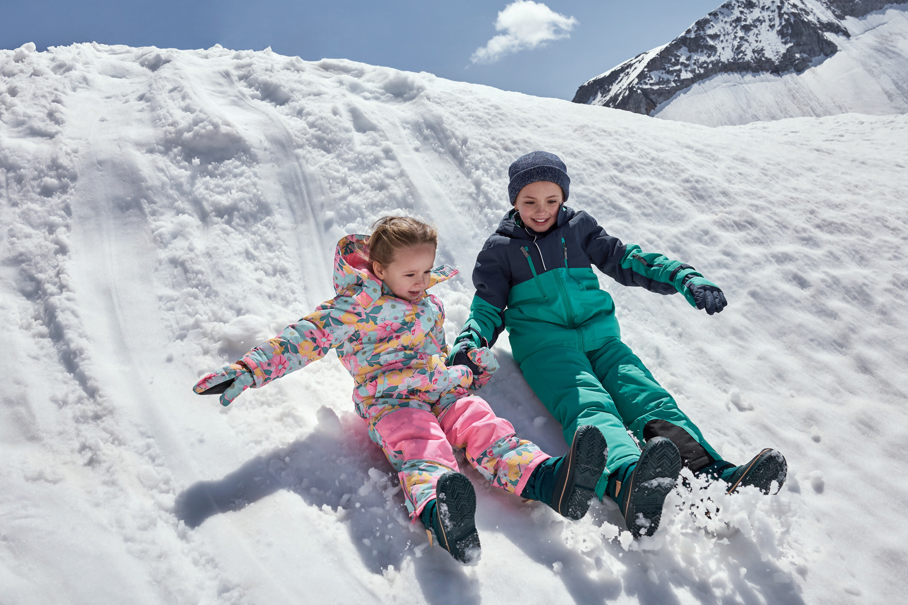 Gyerekek csúsznak le egy havas domboldalon színes téli overallban.
