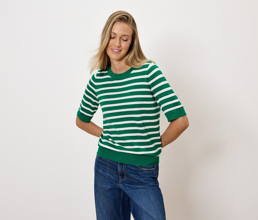 Egy szőke hajú nő zöld-fehér csíkos felsőben és kék farmerben pózol.