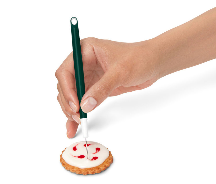 Egy kéz egy kekszet díszít cukormázzal egy zöld dekorációs toll segítségével.