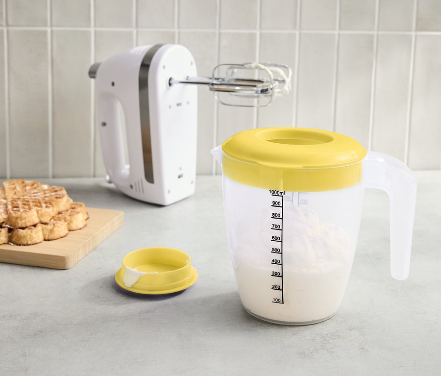A pulton egy fehér mixer, egy sárga fedővel ellátott mérőpohár folyadékkal és egy halom gofri egy deszkán.
