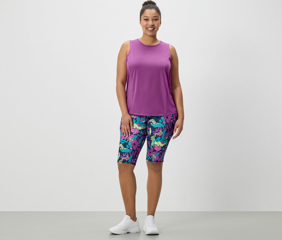 Egy nő áll, nyomott mintás sport rövidnadrágot és egy 2 db-os szettből való türkiz sporttopot visel.