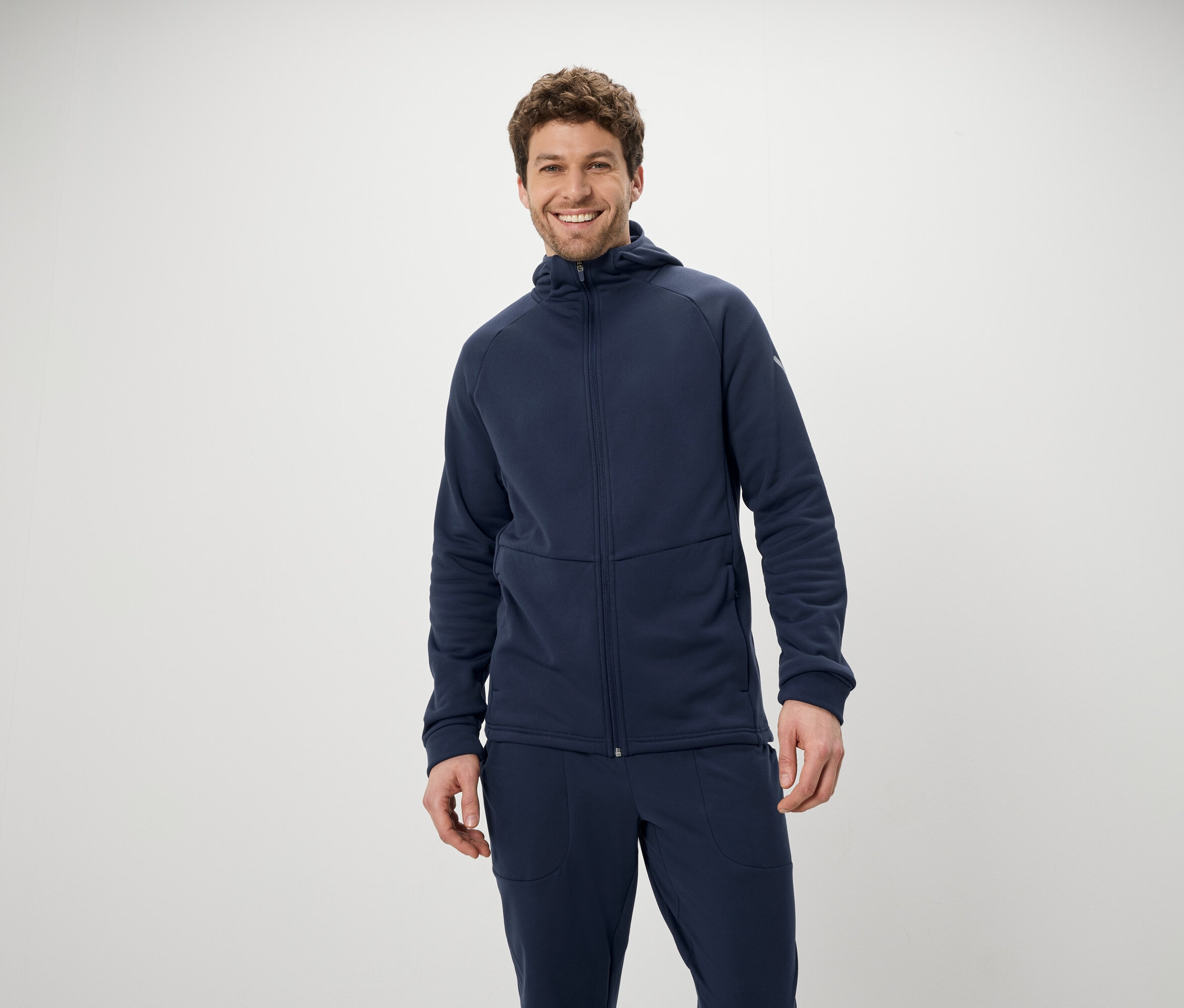 Egy mosolygó férfi kék kapucnis pulóverben és nadrágban pózol fehér háttér előtt.