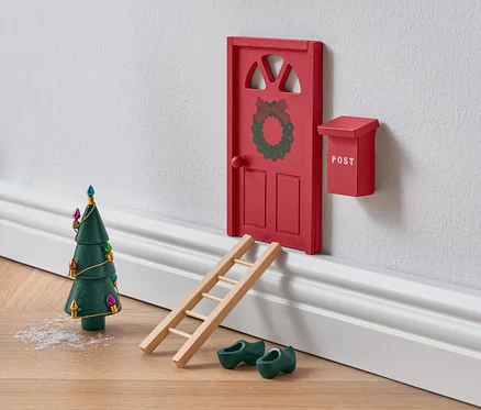 Miniatűr piros ajtó koszorúval, létrával, zöld fapapucsokkal és karácsonyfával.