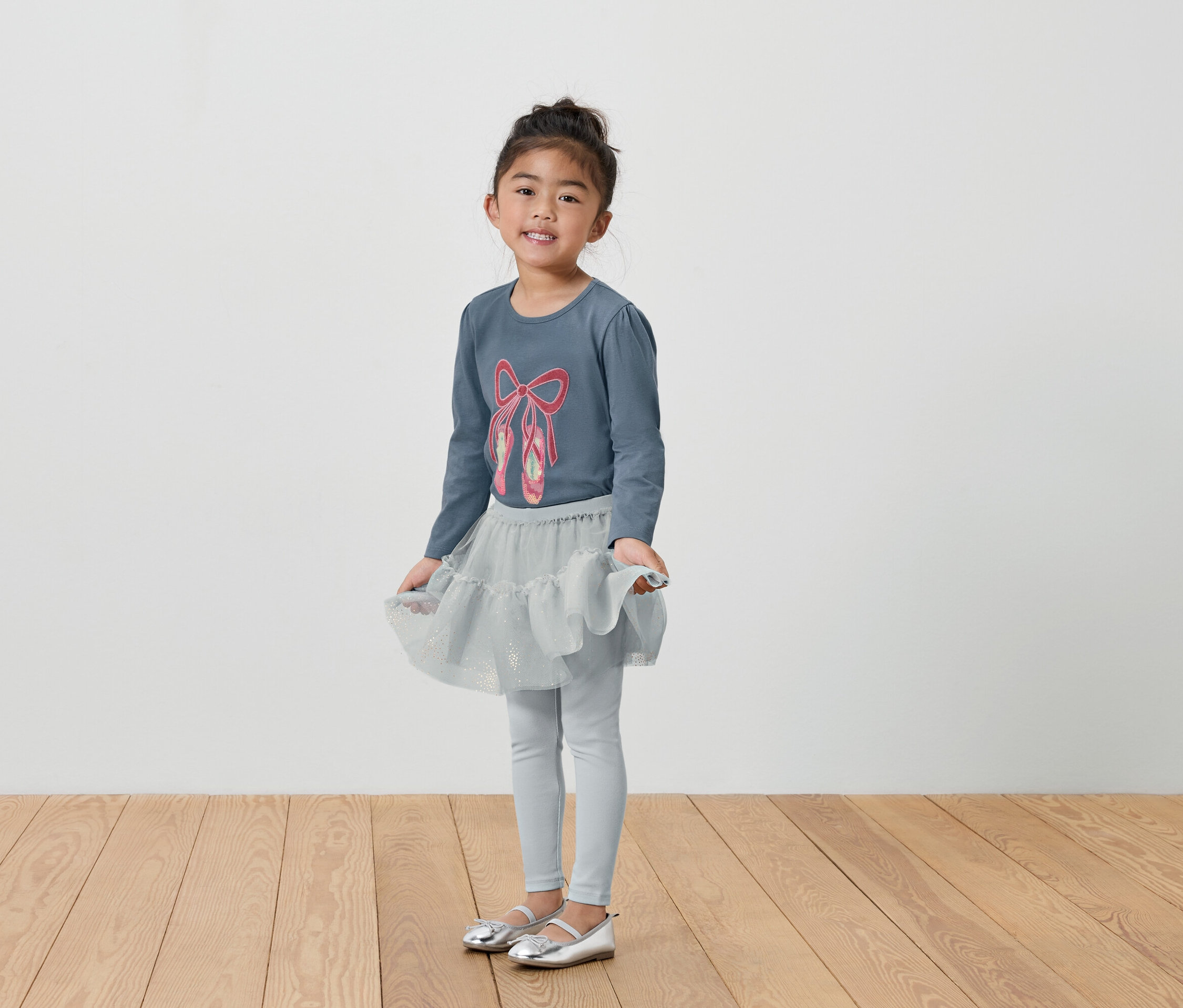 Lány szürke balettruhában, pólóban, szoknyában és leggingsben.