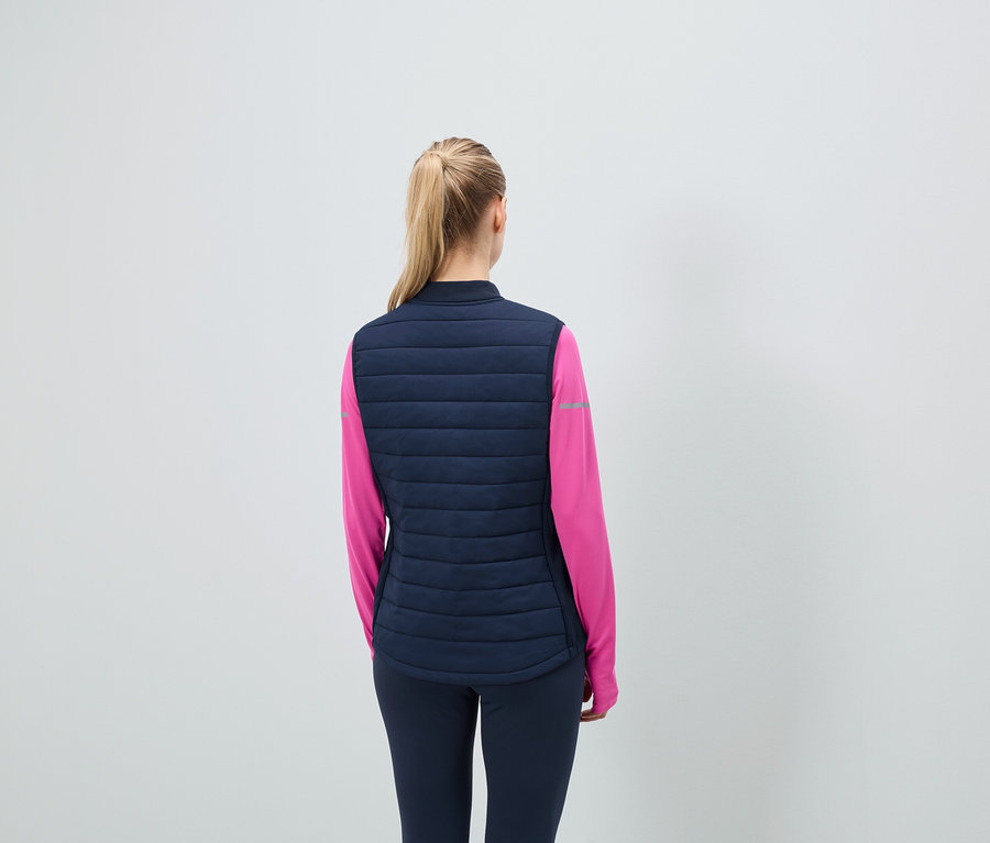 Hátulnézet: szőke hajú, lófarokba kötött nő sötétkék steppelt mellényt és rózsaszín sportfelsőt visel.