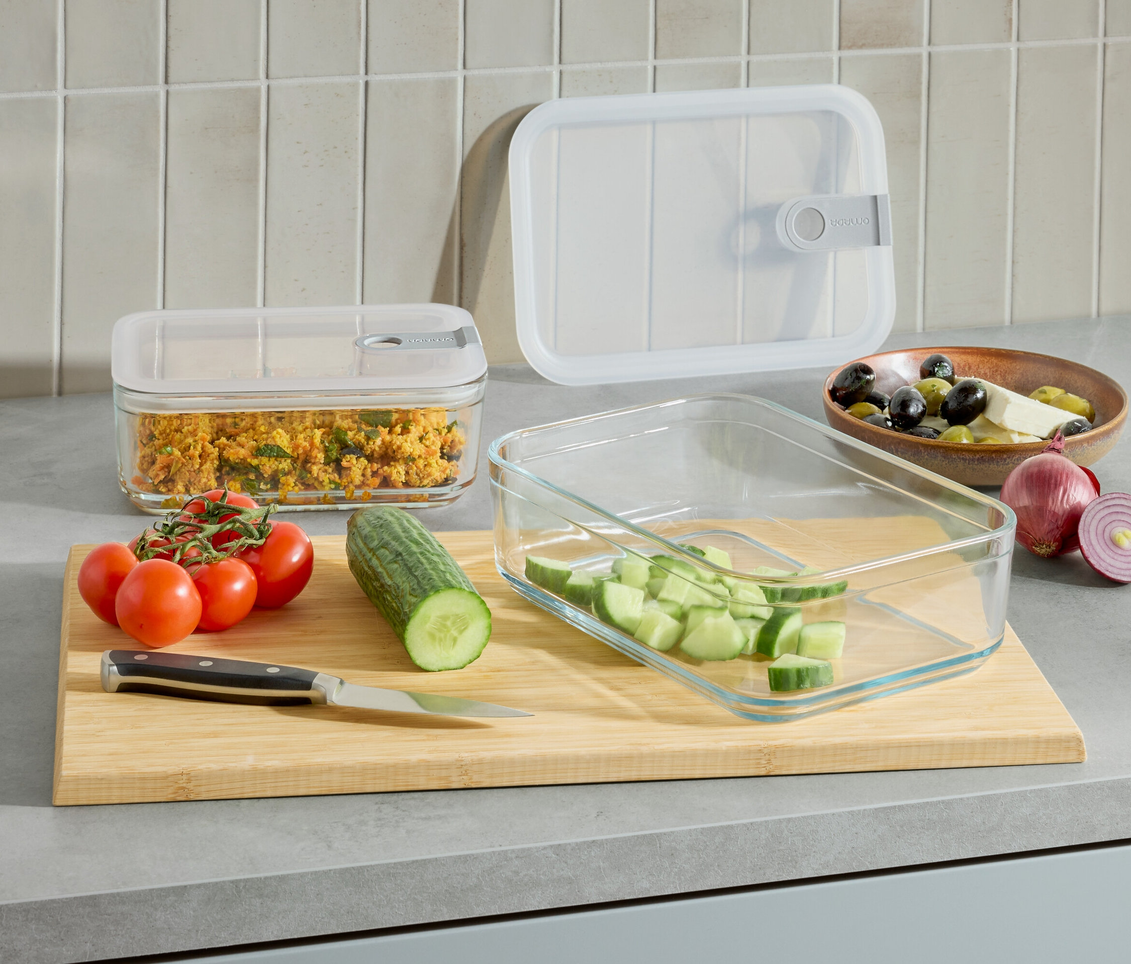 A konyhapulton 2 üveg ételtartó, az egyikben étel, a másikban vágott uborka van. Ezenkívül van paradicsom, uborka, kés és egy tál olívabogyó és sajt.
