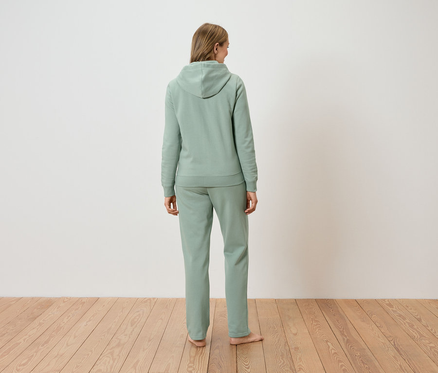 Hátulnézet egy hosszú szőke hajú nőről, aki világoszöld kapucnis pulóvert és hozzáillő nadrágot visel.