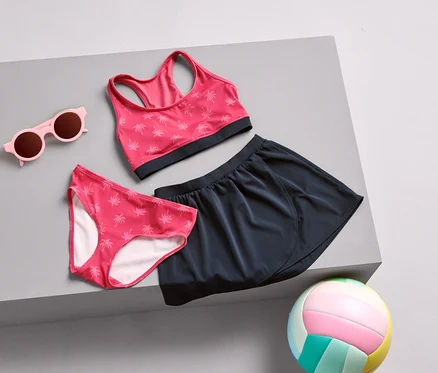 Rózsaszín napszemüveg, pálma mintás bikini alsó, sportmelltartó és tengerészkék szoknya fekszik egy szürke felületen, egy színes röplabdával együtt.