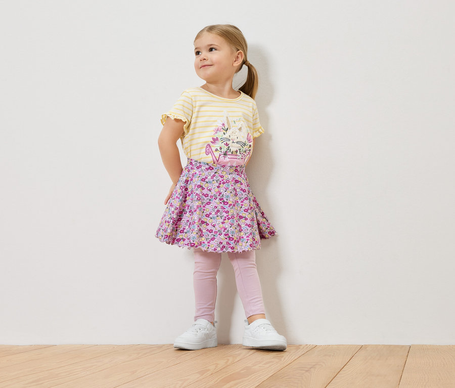 Egy kis szőke lány sárga csíkos, virágmintás pólót, virágos szoknyát, rózsaszín leggingset és fehér tornacipőt visel.