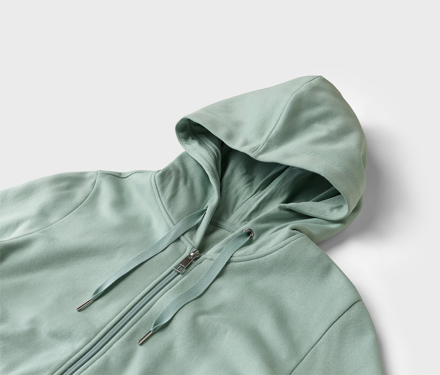 Egy világoszöld, cipzáros kapucnis pulóver közeli képe fehér háttérrel.