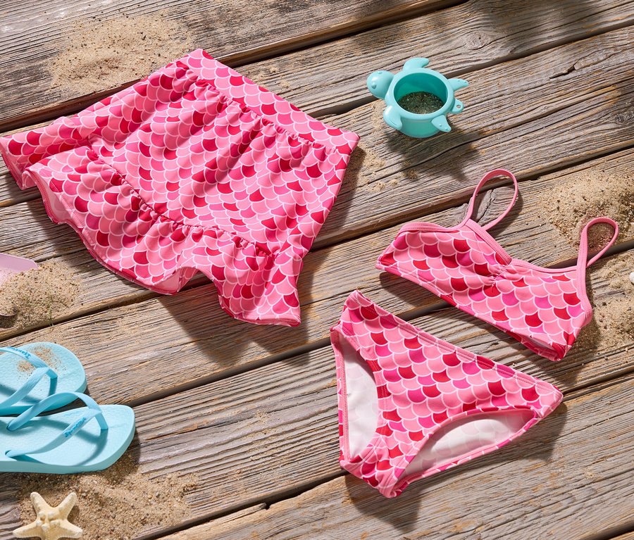 Felülnézet egy rózsaszín sellő bikini szettre, szoknyára, lapátra, flip-flop papucsra és egy teknős homokozó formára a fán.