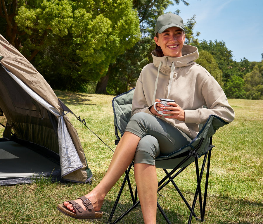 Egy nő ül egy Tambu »ASANA« kempingszéken, egy csészét tart a kezében, és egy 3/4-es outdoor leggingset és egy kapucnis pulóvert visel.