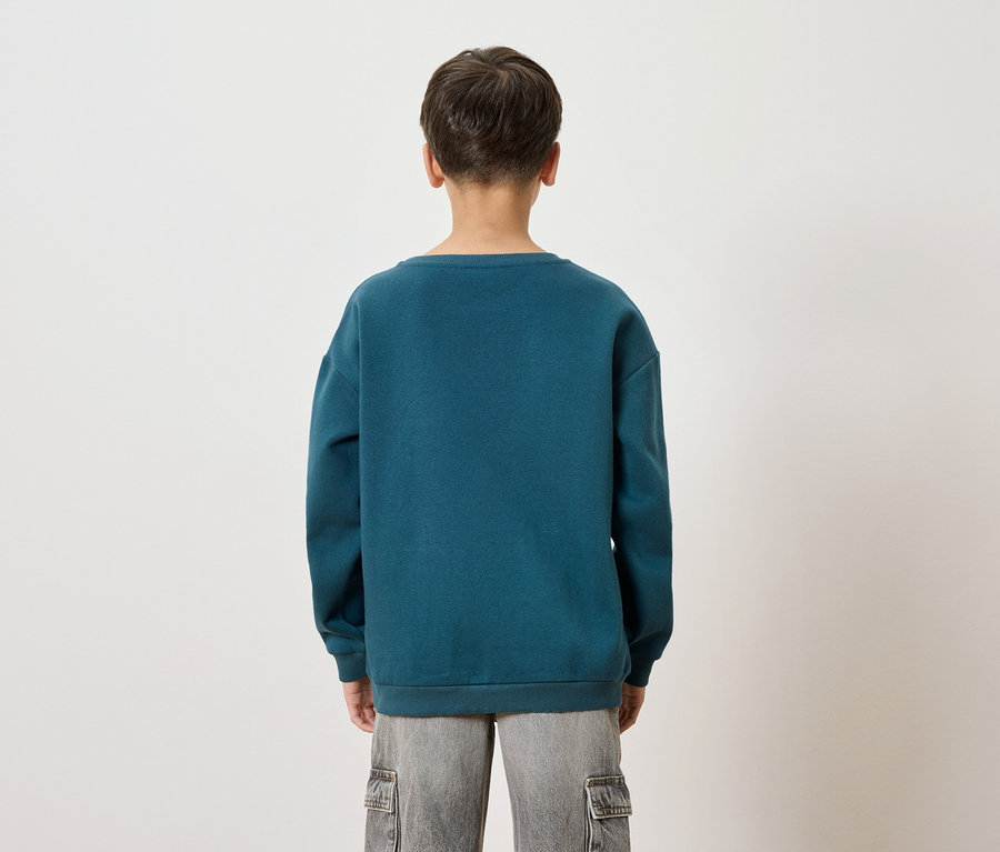 Hátulnézet egy kékeszöld pulóvert és szürke rövidnadrágot viselő gyermekről fehér háttér előtt.