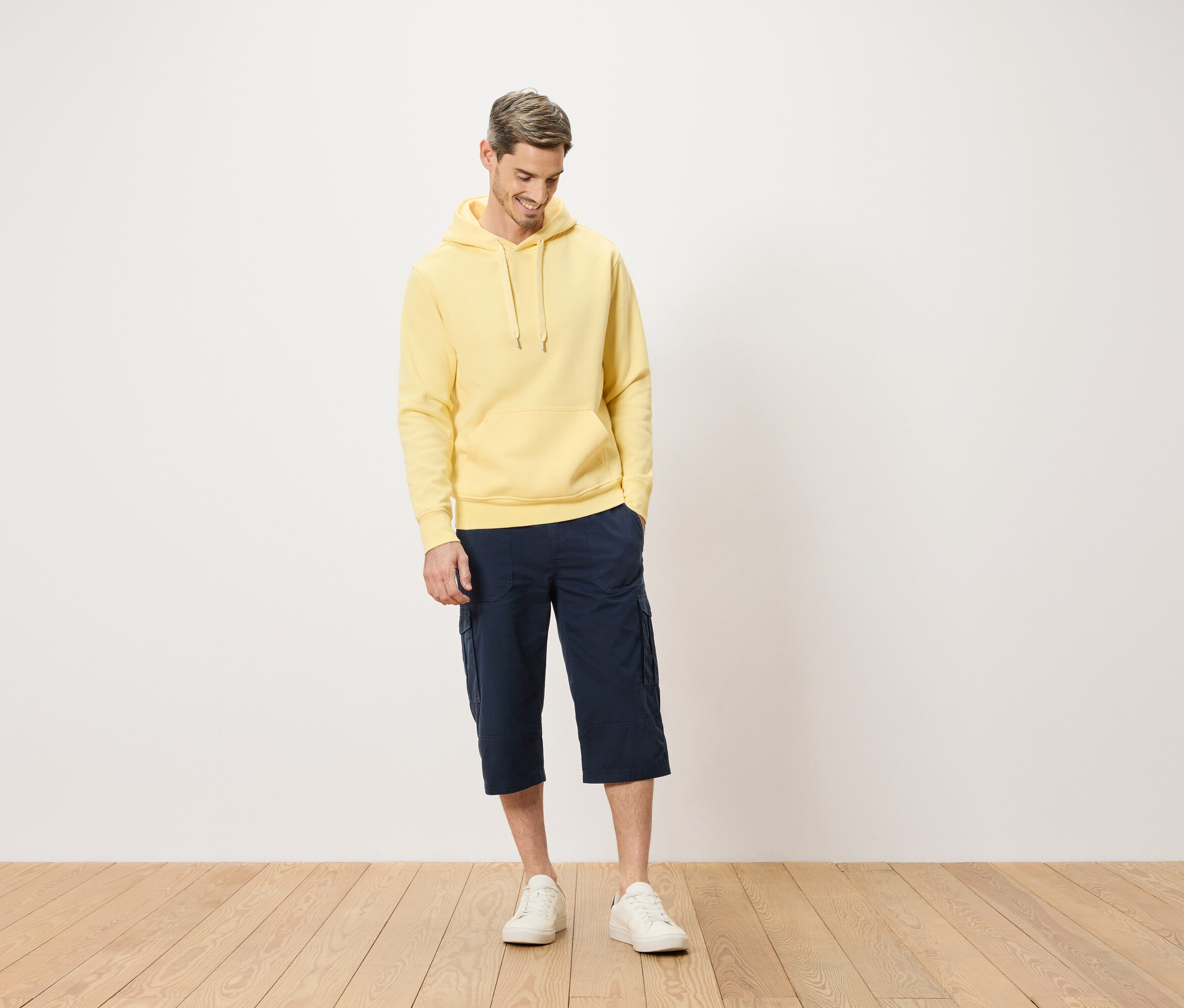 Férfi sárga kapucnis pulóverben és kék cargo rövidnadrágban áll egy fapadlón.
