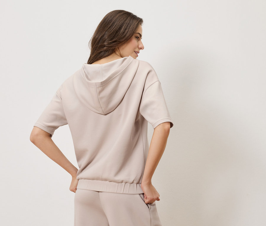 Hátulnézetből egy bézs kapucnis pulóvert és hozzáillő nadrágot viselő nő.