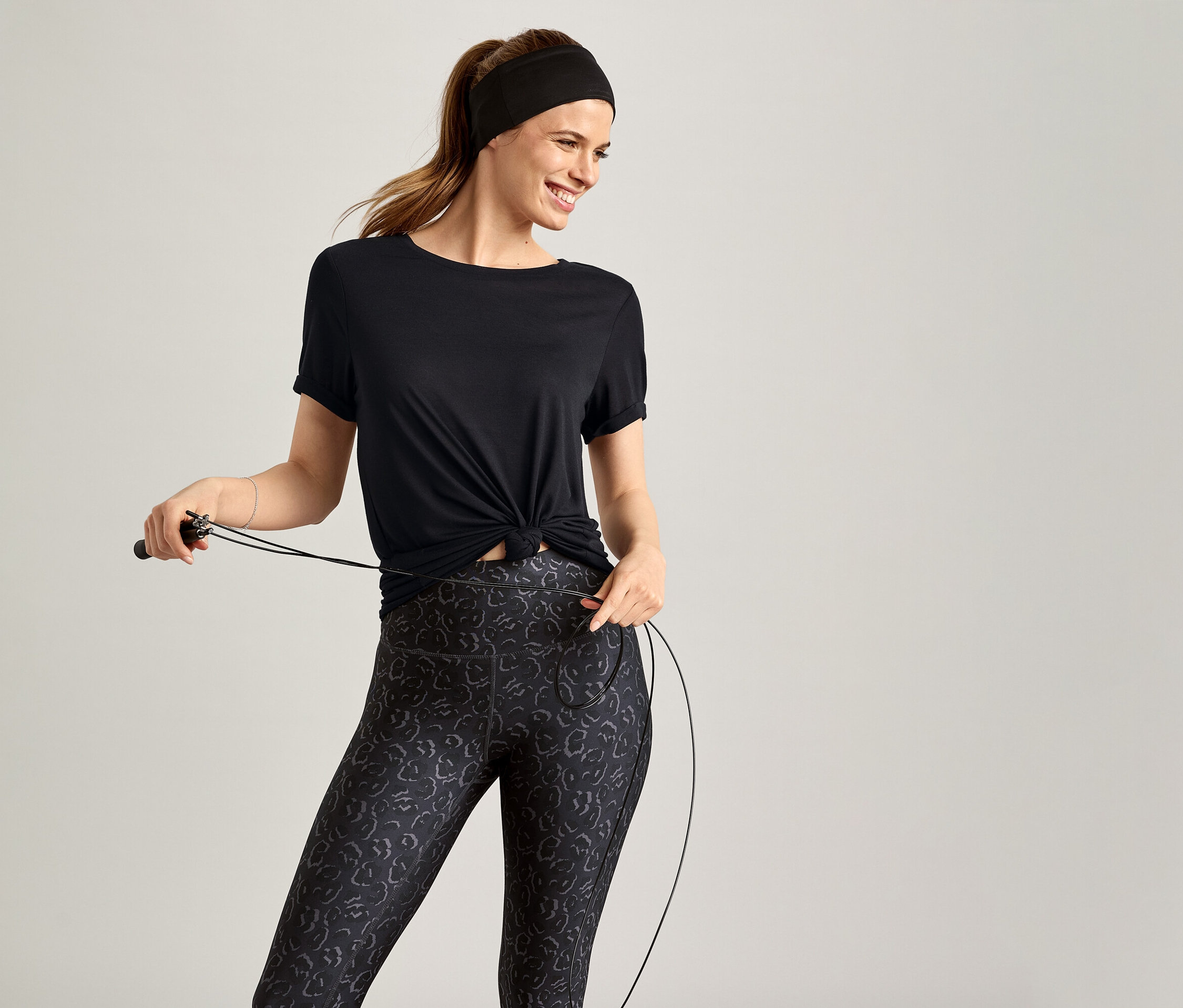 Nő fekete sportfelsőben ugrókötelet tart szürke háttér előtt.