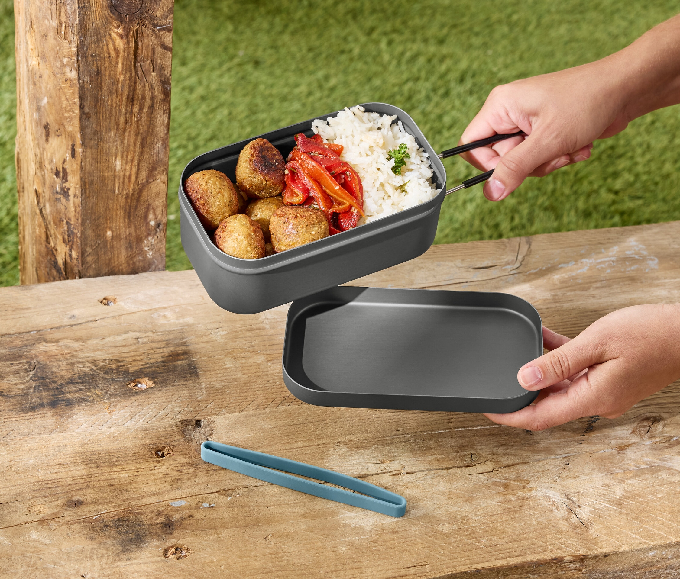 A kezek egy szürke uzsonnásdobozt tartanak rizzsel, húsgombócokkal és paprikával egy szürke fedő felett egy fa asztalon.