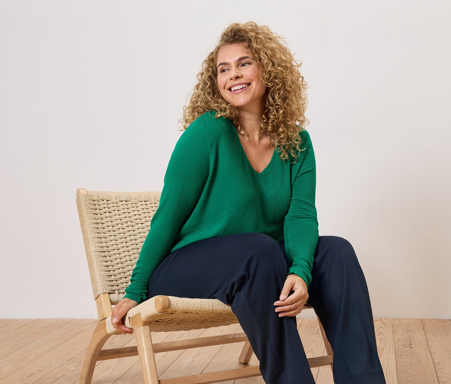 Egy göndör hajú nő ül egy széken, zöld pulóvert és kék nadrágot visel.