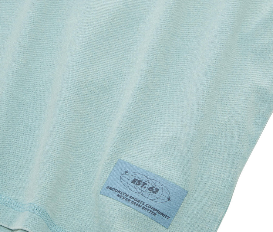 Egy világoskék póló közelképe, rajta egy kék címkével, amelyen a következő felirat olvasható: "EST. 62 BROOKLYN SPORTS COMMUNITY NEVER BEEN BETTER".