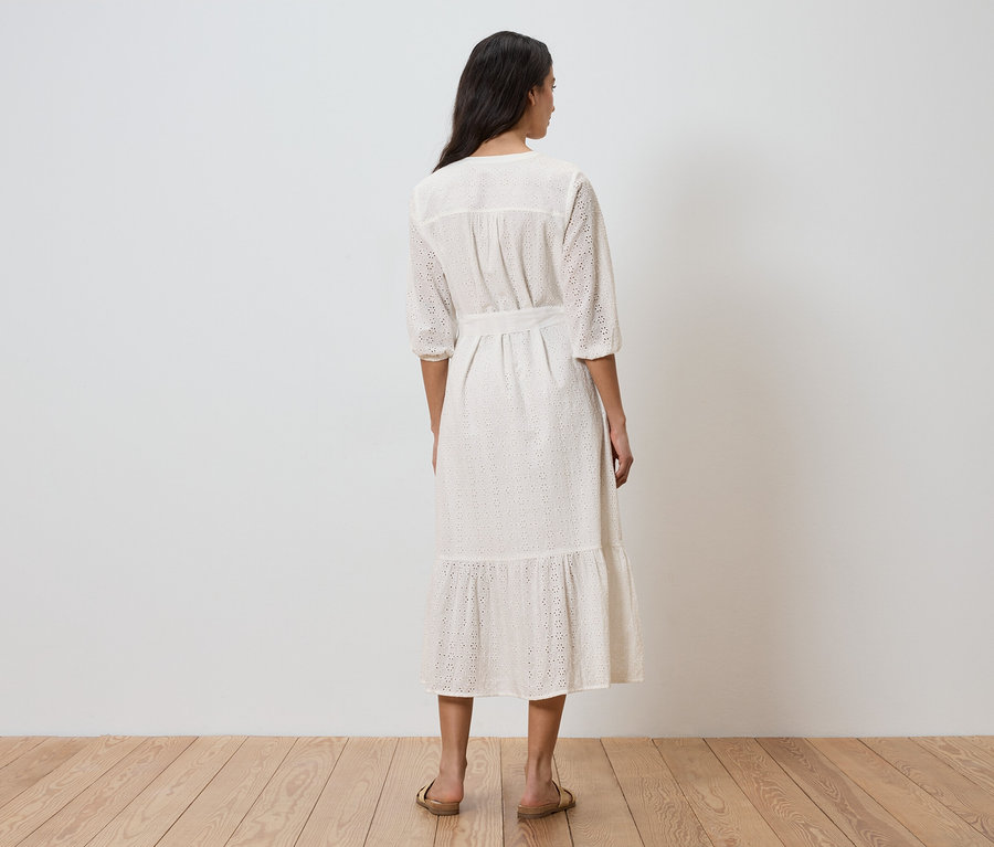 Egy nő háttal áll, fehér ruhát visel övvel és lyukacsos mintával.