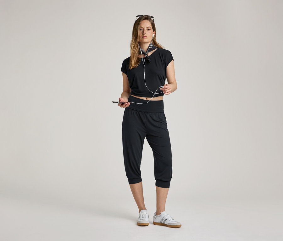 Nő fejhallgatóval és mobiltelefonnal. 3/4-es fekete jóganadrágot, fekete felsőt és tornacipőt visel.