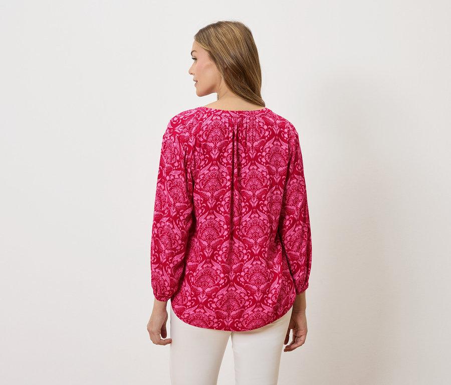 Hátulnézet egy szőke hajú nőről, aki rózsaszín, mintás, hosszú ujjú blúzt és fehér nadrágot visel.