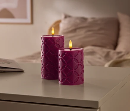 Két geometriai mintás lila LED gyertya világít egy asztalon, mellette egy nyitott könyv, a háttérben pedig egy elmosódott ágy.