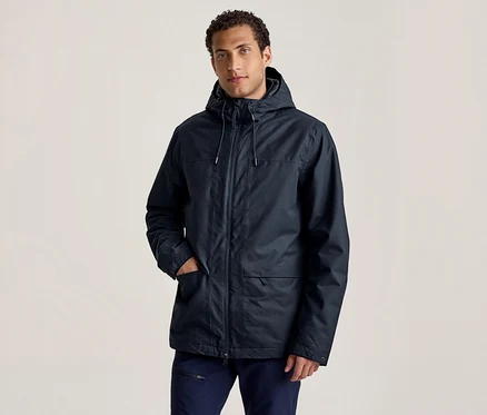 Egy férfi sötétkék kapucnis kabátban és sötétkék nadrágban áll, egyik kezével a kabátzsebében, előre nézve.