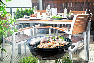 Egy erkély, amelyen egy grill van, rajta pedig húsok. A háttérben pedig egy rusztikus, megterített kerti asztal látható, amely mellett faszékek vannak.