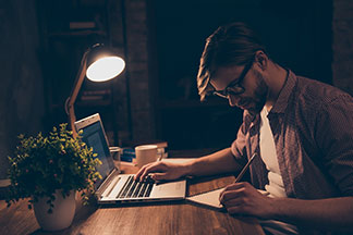 Asztali lámpa az íróasztalon: Egy fiatal ember ül a jól megvilágított, fa íróasztalnál, előtte van egy laptop, éppen egy tollat fog, és ír a jegyzettömbbe - sötét van körülötte.