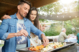 Egy fiatal férfi a grillnél áll, és egy húsnyársat forgat egy grill lapáttal, miközben egy fiatal, hosszúhajú nő átkarolja a vállát, és szintén a grillrácsot nézi.