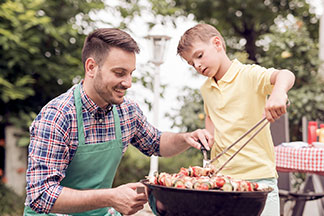Egy zöld kötényes fiatal férfi egy fekete, kerek grill mellett térdel, miközben a fia egy grillcsipesszel húsnyársakat forgat.