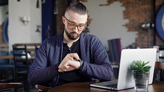 Kardigán az üzletért: Egy szemüveges, sötétkék kardigánt viselő fiatalember egy kávézóban ül a laptopja előtt, és kezeli az okosóráját.