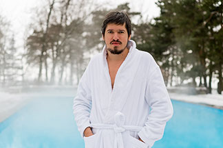 Egy fehér fürdőköpenyes, szakállas férfi áll kint egy gőzölgő medence előtt, kezét a fürdőköpeny zsebében tartva.