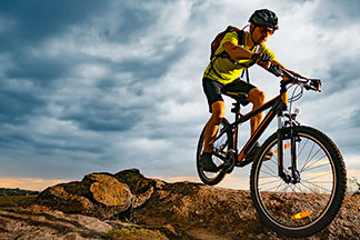 Egy fiatal férfi, aki rövid kerékpáros ruházatként egy rövidujjú kerékpáros mezet és egy rövid kerékpáros nadrágot visel, egy mountain bike-on teker a hegyekben.