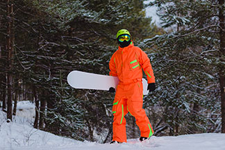Egy narancssárga síoverált viselő snowboardos a havas, téli erdőben gyalogol le a pályáról.
