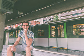 Egy fiatal férfi egy virágos mintával ellátott rövid ujjú ingben és egy rövid chino nadrágban ül a metrómegállóban, egy padon.