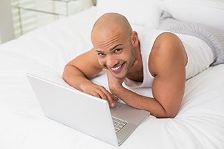 Egy fehér alsóinget és egy hosszú pizsamanadrágot viselő férfi az ágyban fekszik és a laptopján dolgozik.