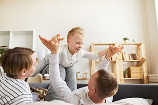 Egy fiatal család otthoni viseletben fekszik az ágyon és az apa a lábain repteti a kisfiát.