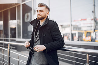 Férfi dzsekik az irodai élethez: Egy fekete rövid kabátot és alatta egy szürke zakót viselő fiatal férfi egy korlátnak támaszkodik és a napszemüvegét a kezében tartja.