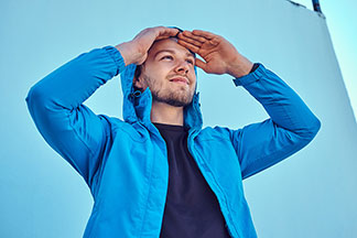 Férfi átmeneti kabátok a szabadba: Egy kék softshell kabátot viselő fiatal férfi a homlokához tartja a kezét és a távolba néz.