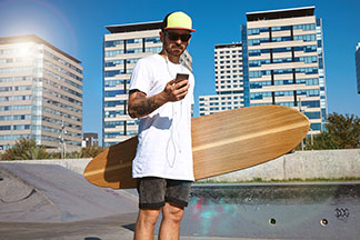 Egy deszkás férfi, napszemüveggel és sapkával a fején egy nagyméretű pólót visel, és longboarddal a kezében egy skate parkban áll és a mobilját nézi.